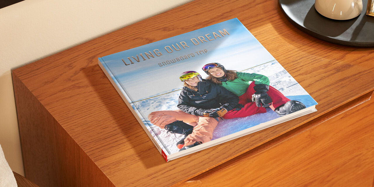 Auf einem Holzschrank liegt ein geschlossenes Fotobuch. Auf dem Cover sieht man ein Foto von zwei Frauen im Schnee. Die Überschrift "Living our Dream" ist in goldenen Buchstaben veredelt.