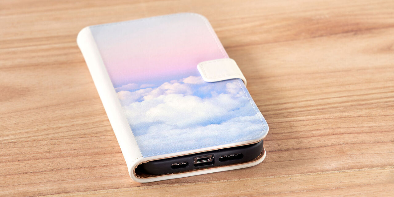Auf einem Tisch liegt ein Smartphone. Es ist die Handyhülle zu sehen, auf der eine Aufnahme von rosafarbenen Wolken zu sehen ist. Bei der Handyhülle handelt es sich um eine Sideflip Tasche aus weissem Leder.